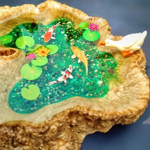 May include: A wooden bowl with a resin pond inside. The pond is filled with green water and has three orange and white koi fish swimming in it. There are also green lily pads and pink flowers floating on the surface. A white duck figurine is perched on the edge of the pond, looking at the fish.