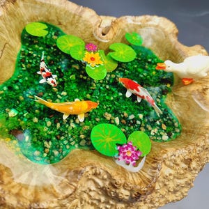 May include: A wooden bowl with a resin pond inside. The pond features green moss, small rocks, and three orange and white goldfish. There are also three green lily pads and two pink and yellow flowers. A white duck figurine is on the edge of the pond.