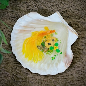 May include: A decorative seashell featuring a painted golden fish, coins, and green details. The fish has a long, flowing tail. The shell is white with a pearlescent sheen and a brown edge. The artwork is set against a brown, textured background.