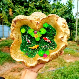 May include: A wooden bowl with a resin inlay depicting a miniature pond scene. The pond is filled with green water, small green lily pads, orange and white fish, and pink flowers. The bowl is a natural wood color and has a rough, textured surface.