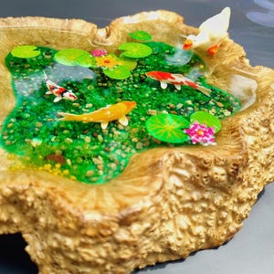 May include: A miniature resin pond scene with a white duck, green lily pads, and colorful fish. The pond is set in a wooden base with a natural bark texture.