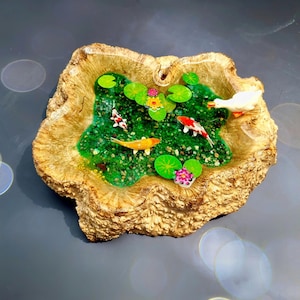 May include: A wooden bowl with a resin pond inside. The pond is filled with green water, small green lily pads, and orange and white fish. There is a white duck figurine swimming in the pond.