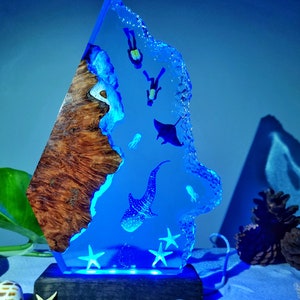 Könnte beinhalten: Eine blaue Harzskulptur eines Walhais, eines Mantarochens und Tauchern, die auf einem Holzsockel mit einem blauen LED-Licht platziert ist. Die Skulptur ist so gestaltet, dass sie wie ein Stück Ozean mit einem Korallenriff auf einer Seite aussieht.