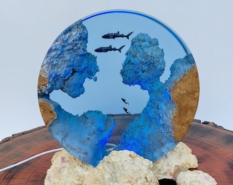 May include: A round resin art piece depicting an underwater scene with blue and gold resin, and black silhouettes of fish swimming. The resin piece is on a white coral base and sits on a wooden surface.