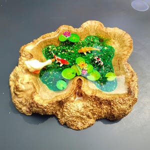 May include: A wooden bowl with a resin pond inside. The pond is filled with green water, green lily pads, and small orange and white fish. There are also pink flowers in the pond.