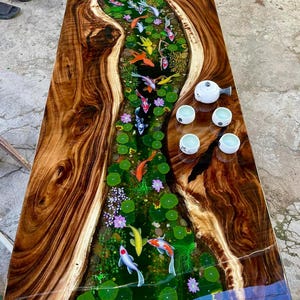 Può includere: Un tavolino rettangolare in legno con un design a forma di fiume. Il centro presenta una resina trasparente con colorate carpe koi, ninfee e fiori. I bordi del tavolo sono in legno marrone scuro e una teiera e tazze bianche sono sulla superficie.