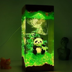 May include: A rectangular resin lamp featuring a panda bear figurine holding bamboo. The lamp has a green and brown landscape design with bamboo stalks and a grassy base. The panda is black and white.