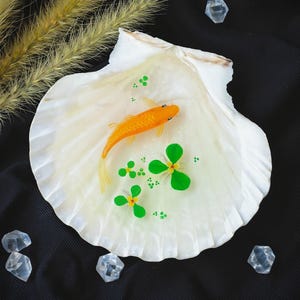 Pode incluir: Uma concha decorativa com um peixe koi laranja em miniatura, nenúfares verdes e pequenos pontos verdes. A concha é branca com um brilho perolado. Cristais transparentes e facetados estão espalhados sobre um fundo preto.