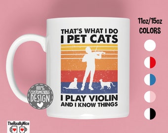 May include: White ceramic mug with a colorful retro design. The mug features a silhouette of a person playing a violin with two cats sitting on either side. The text on the mug reads "That's what I do I pet cats I play violin and I know things".