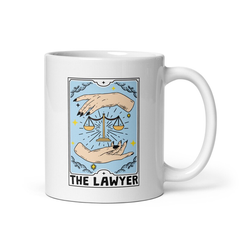 May include: White ceramic mug with a blue and yellow illustration of a tarot card, "The Lawyer", featuring a scale and two hands.