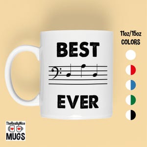 May include: White ceramic mug with the words "BEST EVER" in bold black letters, with a musical staff and notes in between. The mug has a white handle and the text "11oz/15oz COLORS" is visible.