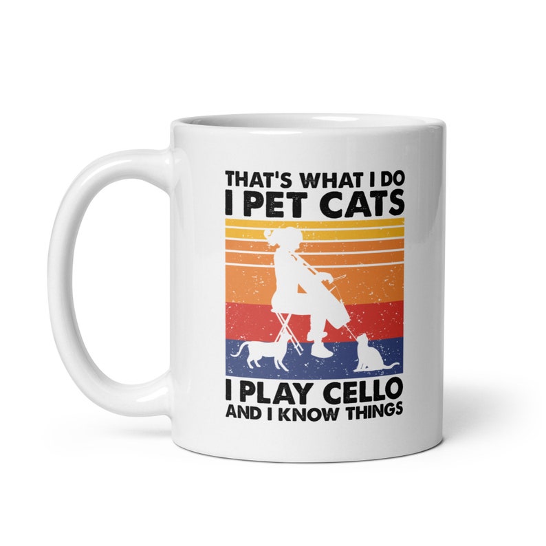 May include: White ceramic mug with a retro-style design featuring a woman playing a cello with two cats. The text reads "That's what I do I pet cats I play cello and I know things."
