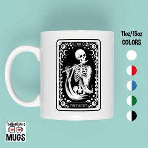 May include: White ceramic mug featuring a black and white tarot card design. The card depicts a skeleton playing a flute, with the text "THE FLUTIST" at the bottom. The mug has a curved handle and the text "11oz/15oz COLORS" is visible.
