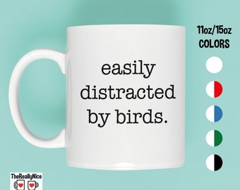 May include: White ceramic mug with the text "easily distracted by birds." The mug is available in 11oz and 15oz sizes and comes in various color options. The mug has a curved handle and is set against a teal background.