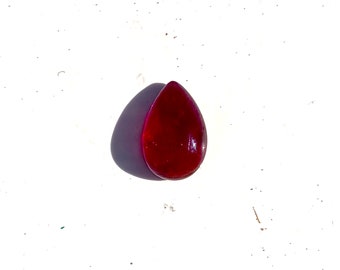 Puede incluir: Una piedra preciosa en forma de lágrima de un color rojo translúcido intenso. La superficie es lisa y pulida, reflejando la luz con un brillo lustroso. La piedra tiene una base redondeada y se estrecha hasta una punta en la parte superior.