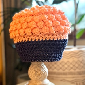 May include: A crocheted hat with a pink and blue color scheme. The top of the hat is made with a bobble stitch, and the bottom is a solid blue.