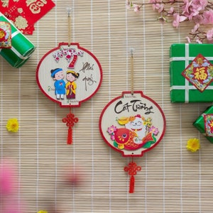 May include: Two round, wooden wall decorations with red trim and gold hanging cords. One features a couple in traditional clothing, the other a cat and fish. Both have red tassels and text in Vietnamese.