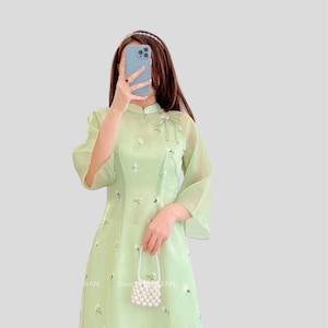 May include: A pale green, semi-transparent dress adorned with floral embroidery. The dress has a mandarin collar, flared sleeves, and a bow at the neckline. A small, beaded purse is held. The dress has the text "Design by HAN" printed on it.