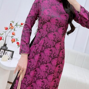 Może przedstawiać: Długa, magenta sukienka z kwiatowym wzorem i długimi rękawami. Sukienka ma kołnierzyk mandaryński i wysoki rozcięcie z boku. Kwiatowy wzór jest ciemnofioletowy. Sukienka jest wykonana z jedwabistego materiału.