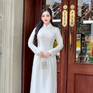 May include: A white, long-sleeved dress with a high neckline and pearl embellishments. The dress is floor-length and is paired with a small, beaded purse. The background features a wooden door with gold accents.