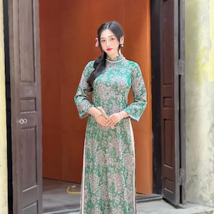 May include: A green floral patterned dress with long sleeves and a high collar. The dress has a pink hem and is worn by a person standing in front of a doorway. The dress is a traditional style.