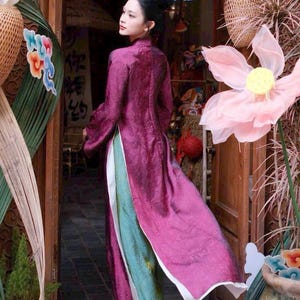 May include: A woman wearing a long, flowing, magenta dress with a high collar and long sleeves. The dress has a white trim and is layered over a teal and green garment. The background includes decorative floral elements and a wooden doorway.