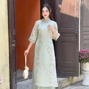 Könnte beinhalten: Ein hellblaues Kleid mit Blumenmuster und eine passende Hose. Das Kleid hat einen Stehkragen und Dreiviertelärmel. Die Frau hält eine weiße Tasche.