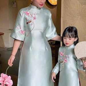 Pode incluir: Dois vestidos verde claro a condizer com bordados florais cor-de-rosa. Os vestidos têm gola mandarim e são compridos até ao chão. Um é usado por um adulto, o outro por uma criança. O adulto segura orquídeas cor-de-rosa.