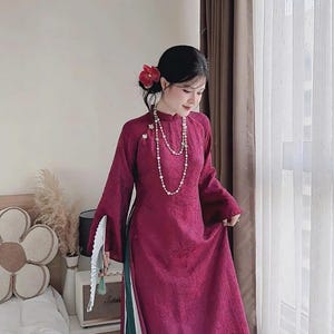 May include: A long, magenta-colored dress with long sleeves and a high collar. The dress has a split on the side, revealing green pants underneath. A white fan is held in the hand. A pearl necklace and a flower in the hair complete the look.