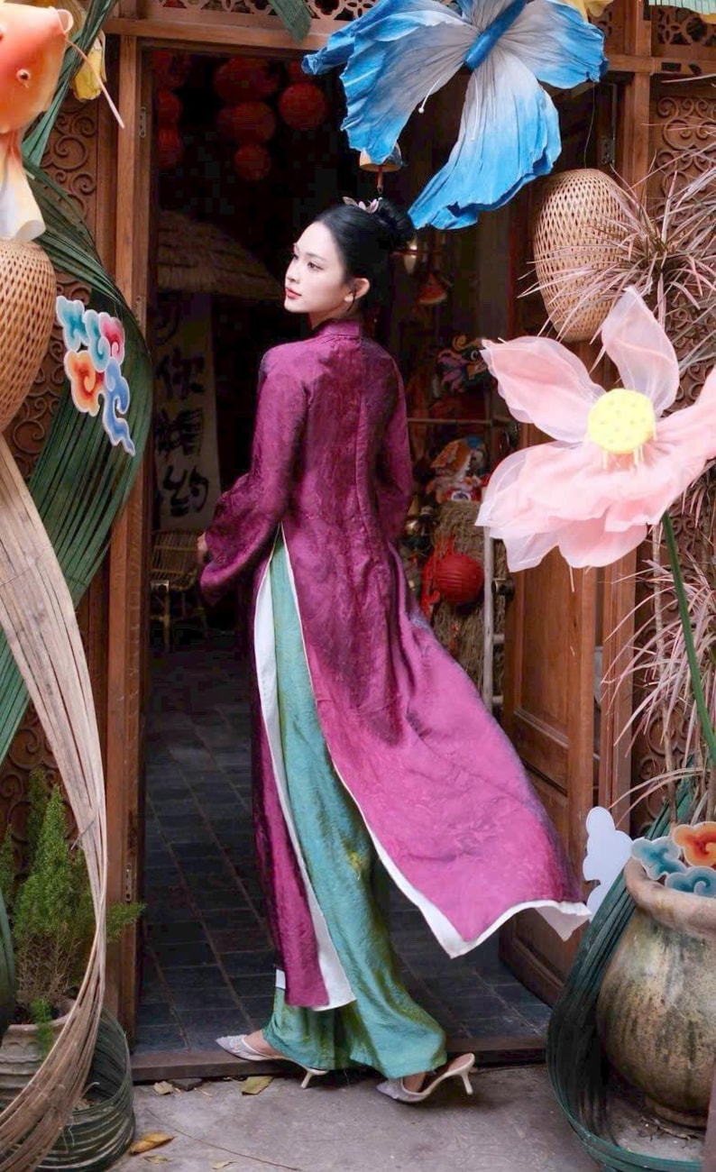 May include: A woman wearing a long, flowing, magenta dress with a high collar and long sleeves. The dress has a white trim and is layered over a teal and green garment. The background includes decorative floral elements and a wooden doorway.