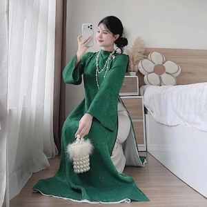 May include: A long, emerald green dress with a floral pattern and wide sleeves. The dress is paired with white pants and a pearl necklace. A small, white beaded purse with a fluffy trim is held in the hand.