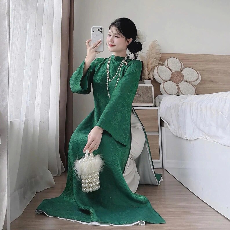May include: A long, emerald green dress with a floral pattern and wide sleeves. The dress is paired with white pants and a pearl necklace. A small, white beaded purse with a fluffy trim is held in the hand.