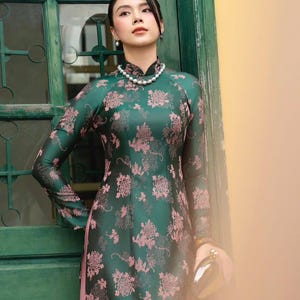 May include: A long-sleeved, emerald green dress with a floral pattern in shades of pink. The dress features a mandarin collar and a pearl necklace. A small, silver clutch is held in the hand. The background includes a green door and a yellow wall.