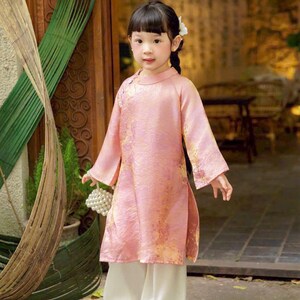 May include: A child wearing a long-sleeved, light pink tunic with a subtle gold floral pattern, featuring a high collar and side slits, paired with loose-fitting white trousers. The outfit is completed with dark shoes and a small white beaded bag.