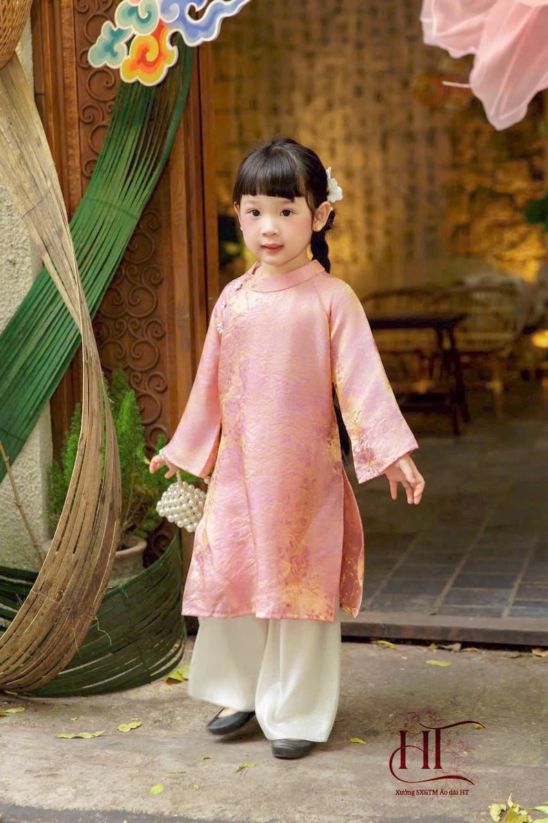 May include: A child wearing a long-sleeved, light pink tunic with a subtle gold floral pattern, featuring a high collar and side slits, paired with loose-fitting white trousers. The outfit is completed with dark shoes and a small white beaded bag.