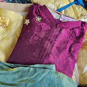 May include: Assortment of colorful, folded garments, including pink, yellow, and purple tops with decorative gold-colored buttons. Some items are in clear plastic packaging. The garments appear to be made of a silky fabric.