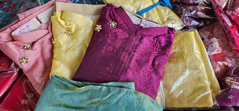May include: Assortment of colorful, folded garments, including pink, yellow, and purple tops with decorative gold-colored buttons. Some items are in clear plastic packaging. The garments appear to be made of a silky fabric.