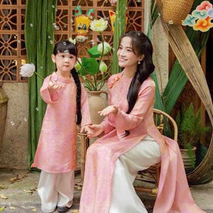 May include: A matching set of two pink long-sleeved tunics with a subtle tonal pattern, each paired with loose-fitting white trousers. The tunics feature a high collar and side slits, worn by two individuals in a coordinated traditional style.