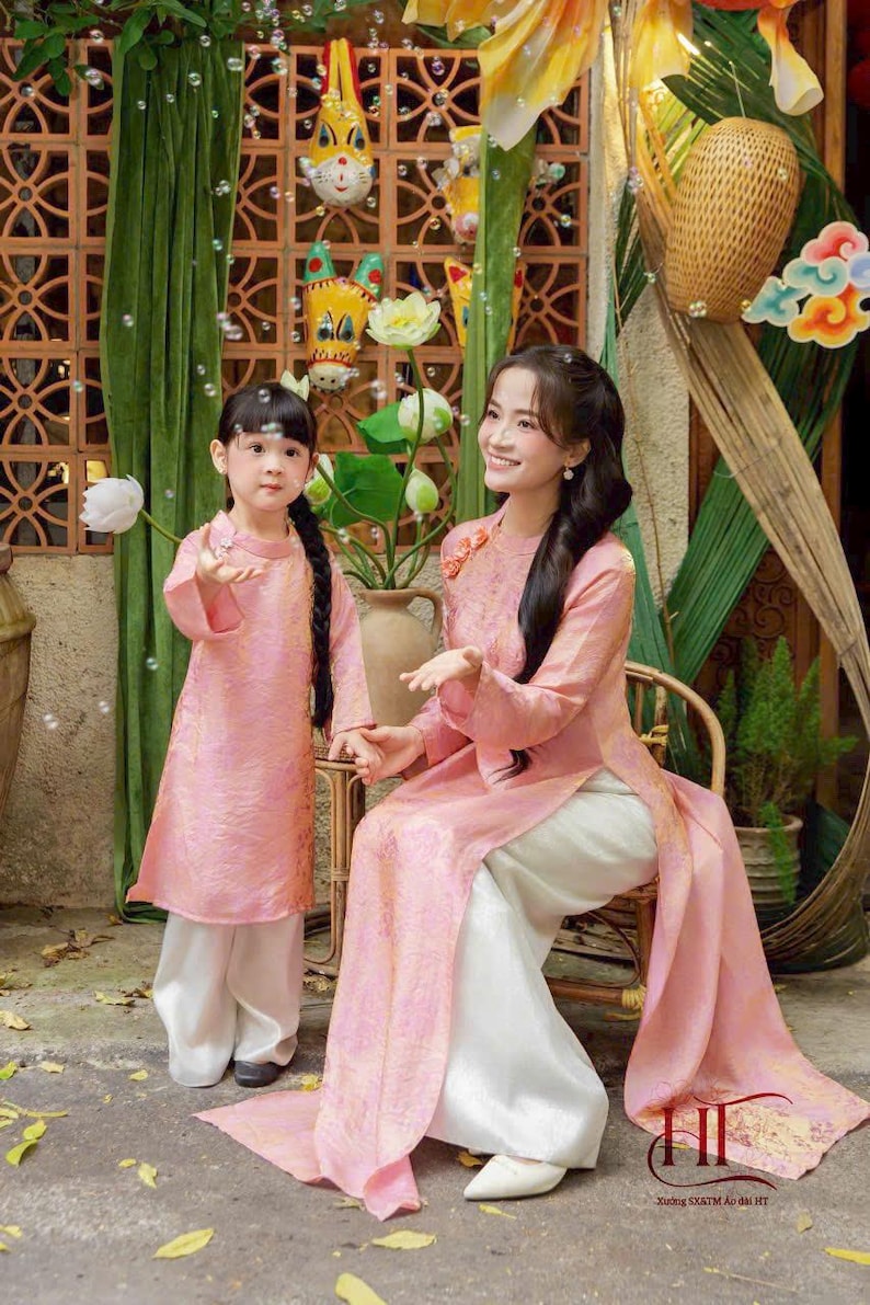 May include: A matching set of two pink long-sleeved tunics with a subtle tonal pattern, each paired with loose-fitting white trousers. The tunics feature a high collar and side slits, worn by two individuals in a coordinated traditional style.