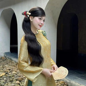 May include: A woman in a gold-colored traditional dress with long black hair styled with decorative hair accessories. She holds a fan and smiles. The dress has long sleeves and a green trim. The background features an arched structure.
