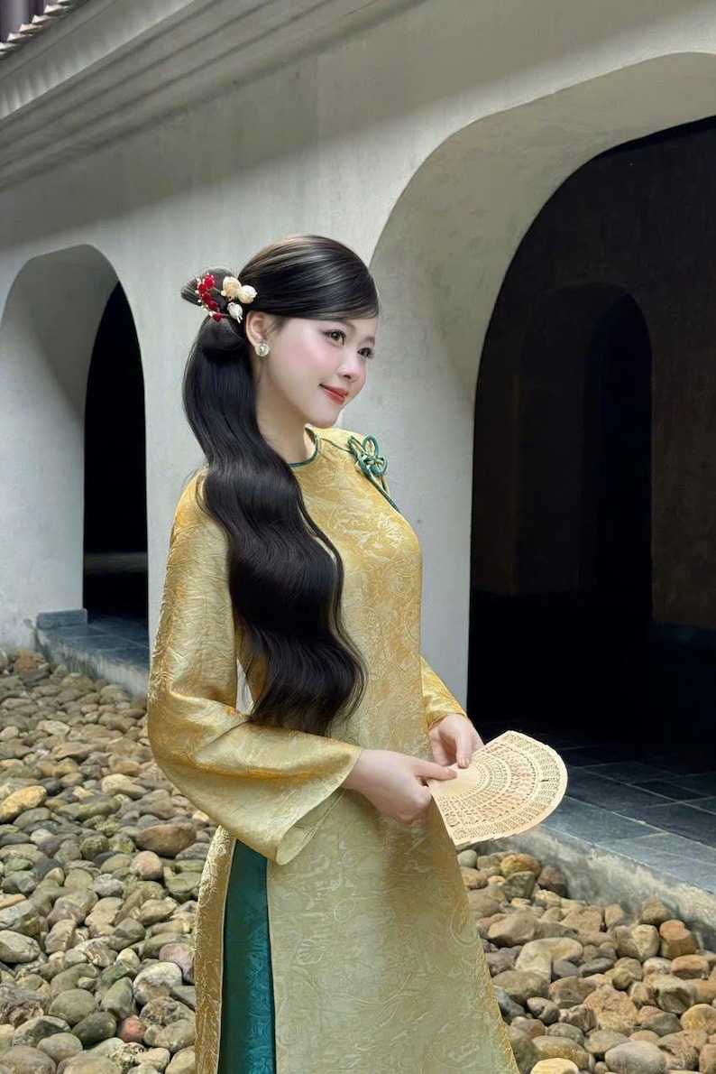 May include: A woman in a gold-colored traditional dress with long black hair styled with decorative hair accessories. She holds a fan and smiles. The dress has long sleeves and a green trim. The background features an arched structure.