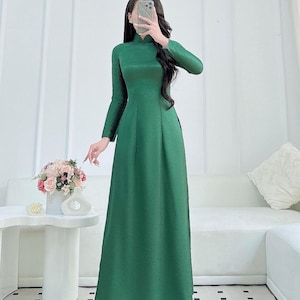 Könnte beinhalten: Ein langes, smaragdgrünes Kleid mit langen Ärmeln und Stehkragen. Dazu wird eine passende Hose mit weitem Bein getragen. Das Outfit besteht aus einem glänzenden, seidigen Material. Die Person hält ein Telefon vor ihr Gesicht.