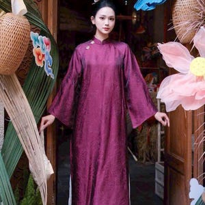 May include: A woman wearing a long, flowing, plum-colored dress with wide sleeves and a mandarin collar. The dress has a subtle floral pattern. The background includes decorative floral and butterfly accents, and a wooden doorway.