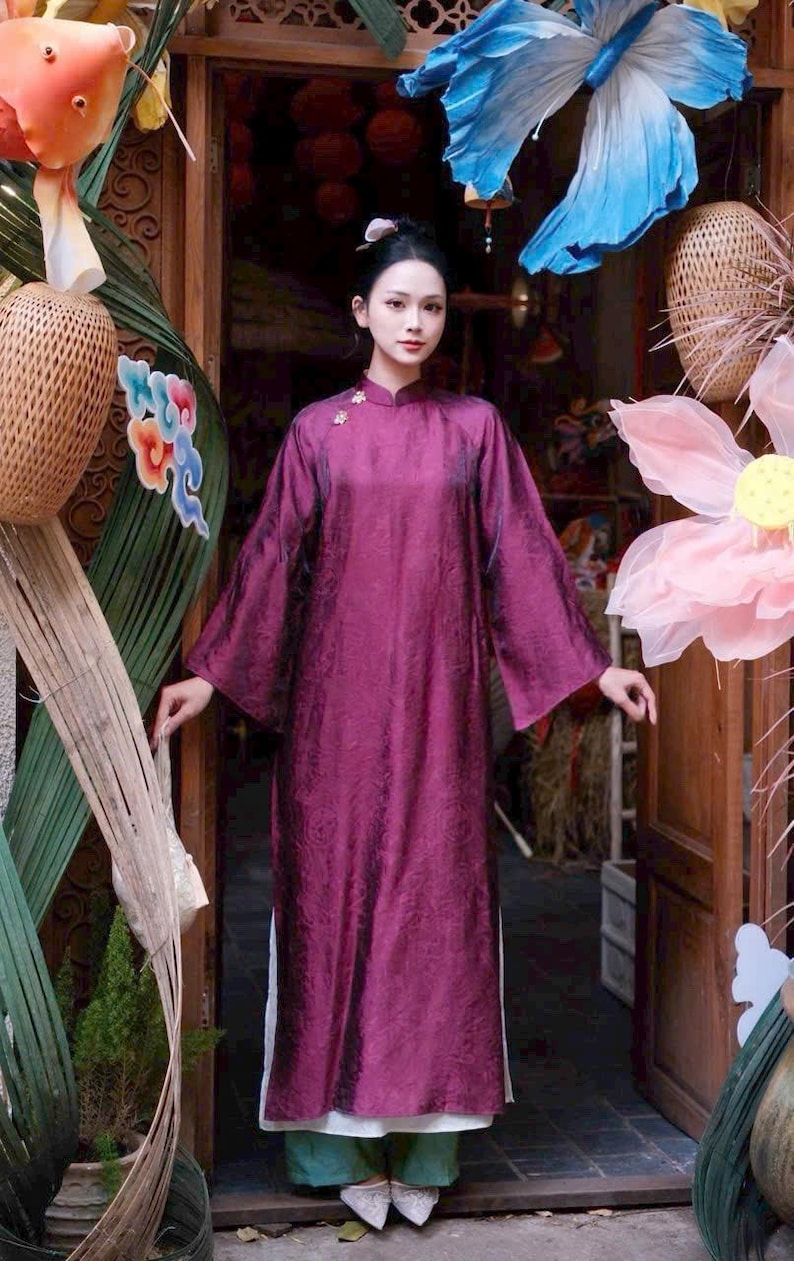May include: A woman wearing a long, flowing, plum-colored dress with wide sleeves and a mandarin collar. The dress has a subtle floral pattern. The background includes decorative floral and butterfly accents, and a wooden doorway.