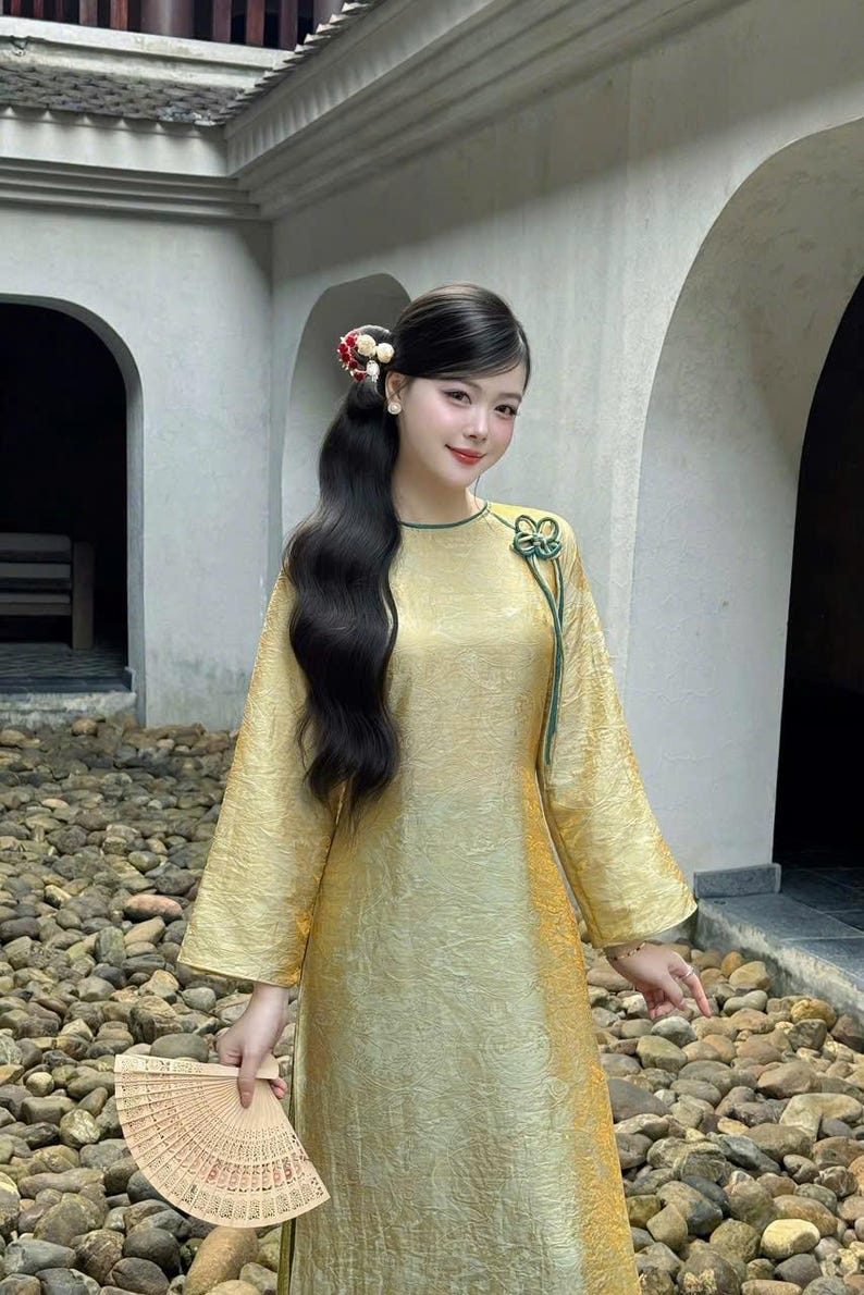 May include: A woman wearing a gold-colored dress with long sleeves and a green trim. She has long black hair and is holding a decorative fan. The dress has a floral design on the front.