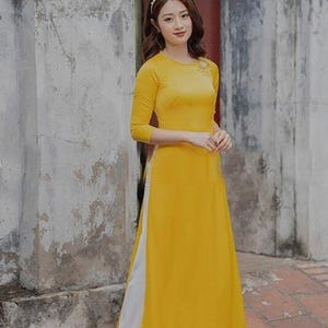 May include: A bright yellow long-sleeved tunic dress featuring a round neckline with subtle decorative embroidery on the left shoulder, worn over loose-fitting white trousers. The ensemble is paired with simple white flat shoes and a pearl headband.