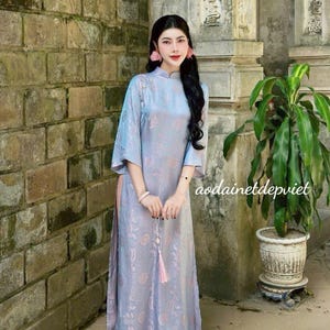 May include: A light blue and pink floral patterned dress with a high collar and three-quarter sleeves. The dress is worn with pink trousers. The text "avdainetdepviet" is visible on the right side of the image.