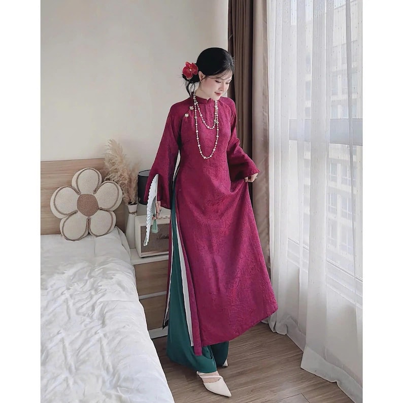 May include: A long, magenta-colored dress with long sleeves and a high collar. The dress has a split on the side, revealing green pants underneath. A white fan is held in the hand. A pearl necklace and a flower in the hair complete the look.