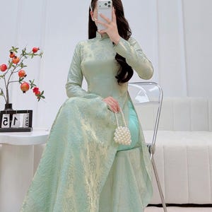 May include: A light green, long-sleeved dress with a high collar and floral pattern. The dress has a flowing skirt and is paired with a small, white beaded purse. The person is seated on a clear chair.