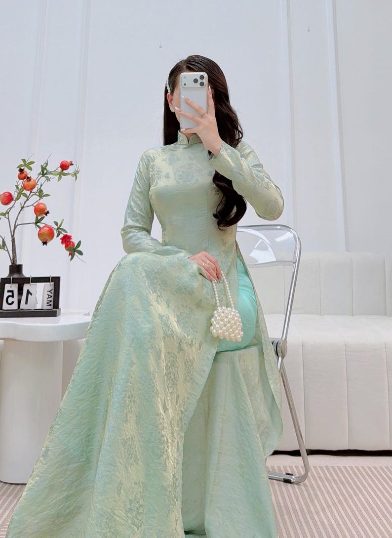 May include: A light green, long-sleeved dress with a high collar and floral pattern. The dress has a flowing skirt and is paired with a small, white beaded purse. The person is seated on a clear chair.